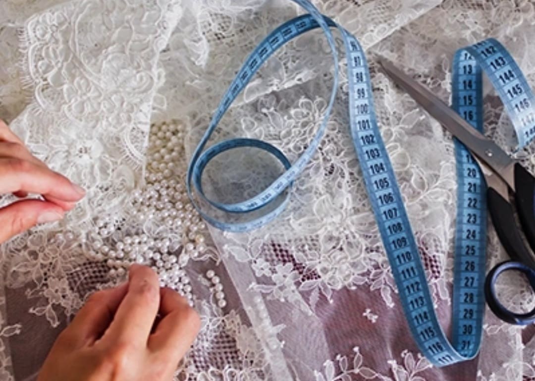 Hands sewing pearls onto white lace fabric, with a blue measuring tape and scissors nearby.