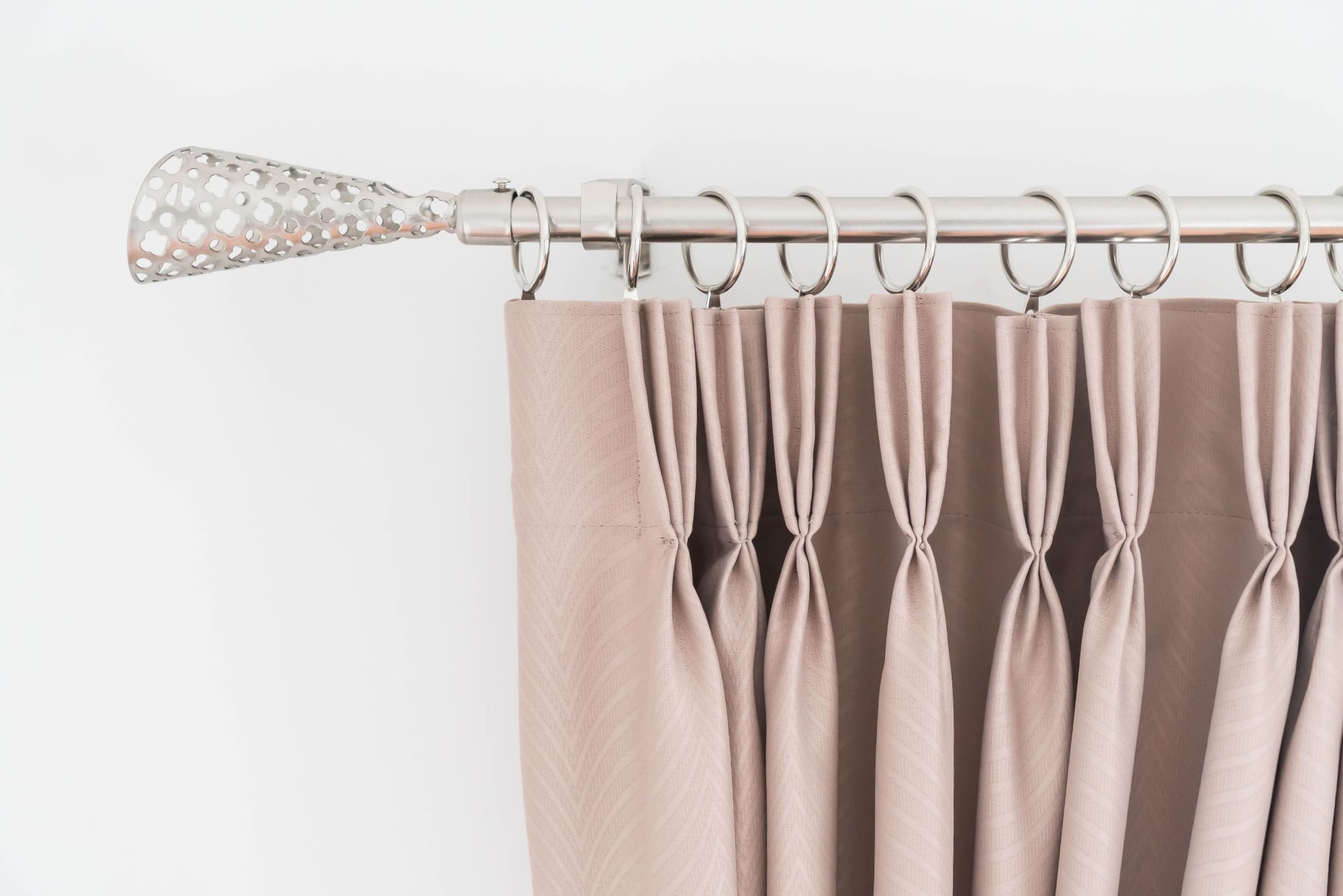 A close-up of beige pleated curtains hanging from a silver curtain rod with decorative finial and metal rings against a white wall.