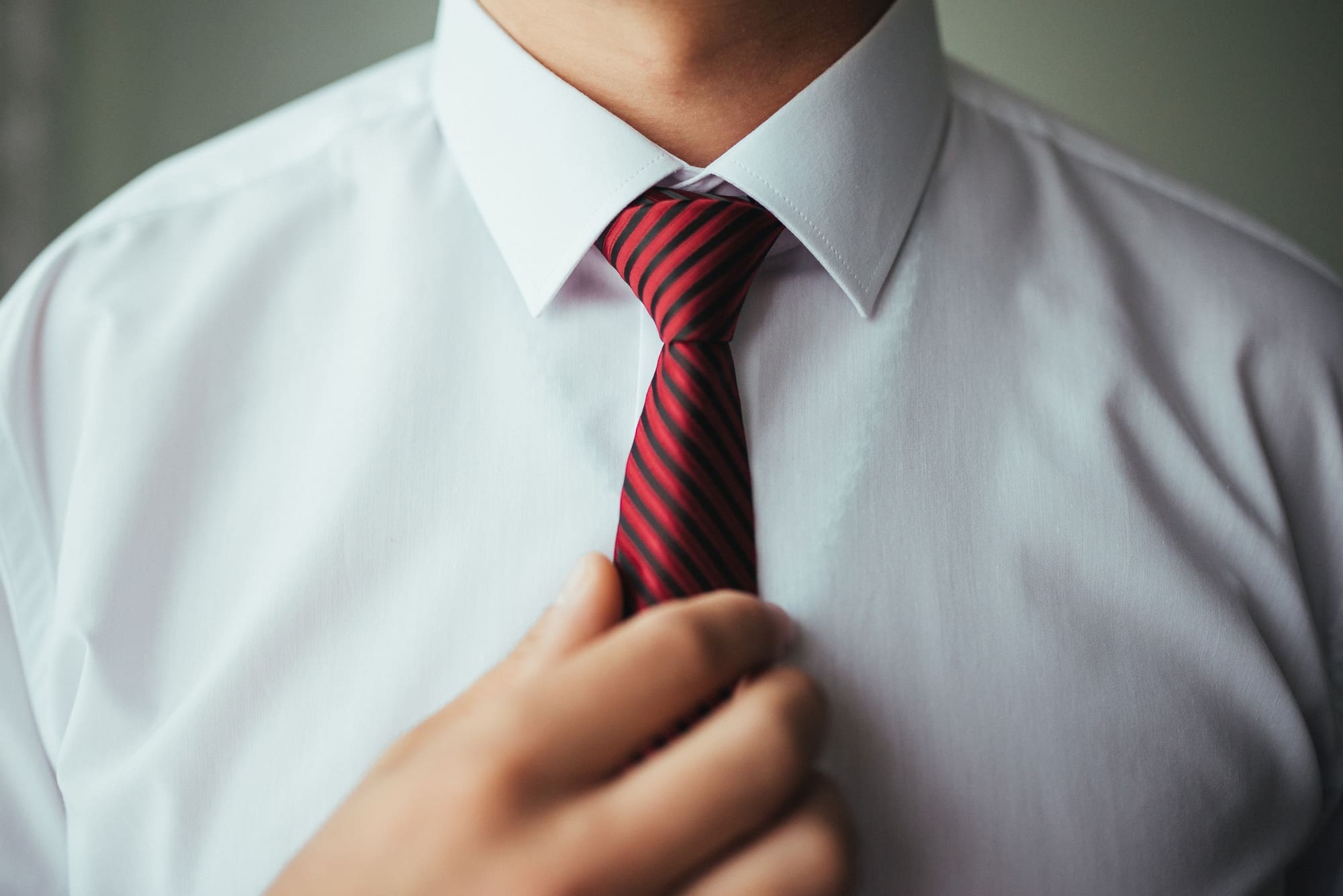 A person wearing a white dress shirt adjusts a red and black striped necktie.