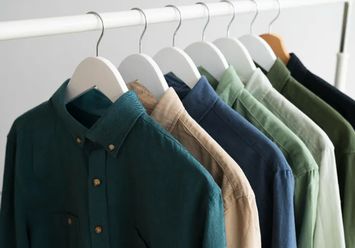 Six collared, long-sleeve shirts in various shades of blue, green, and beige hang neatly on white hangers from a white clothing rack.