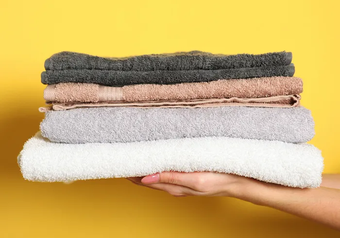 A hand holds a neatly folded stack of four towels in different colors—dark gray, beige, light gray, and white—against a yellow background.
