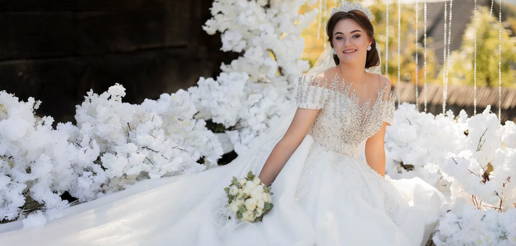 A bride in a white wedding dress sits among white flowers, holding a bouquet and smiling at the camera.