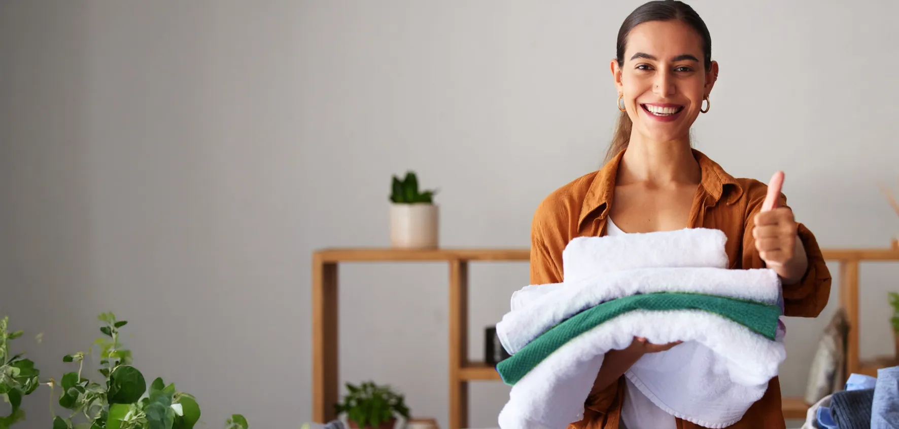 A person stands indoors holding a stack of clean, folded towels and gives a thumbs up, smiling. Shelves with plants and decor are in the background.