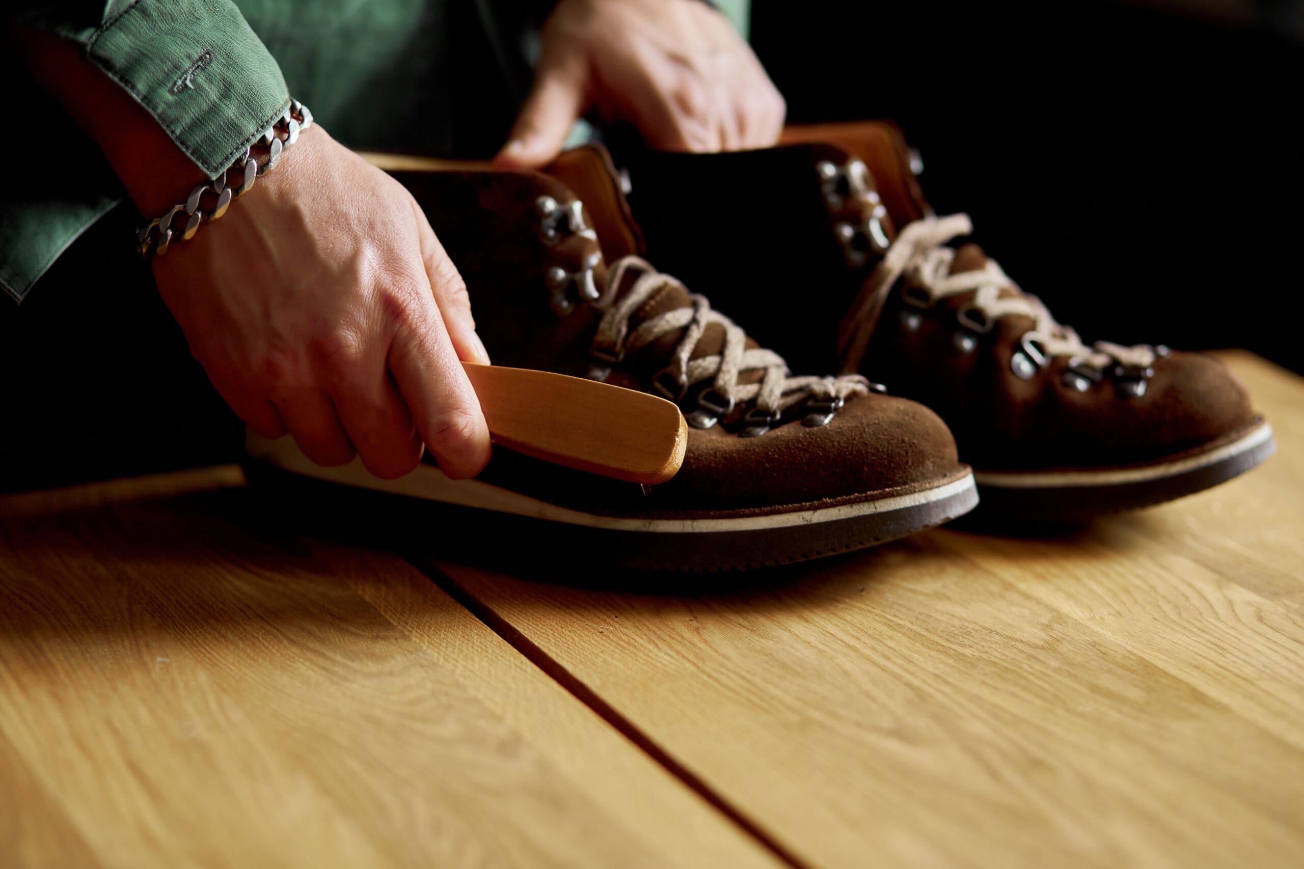 A person uses a brush to clean a pair of brown suede hiking boots on a wooden surface.