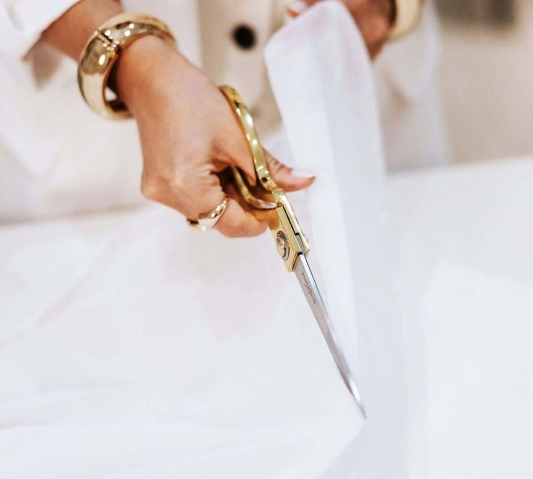 A person wearing gold jewelry uses gold scissors to cut white fabric on a table.