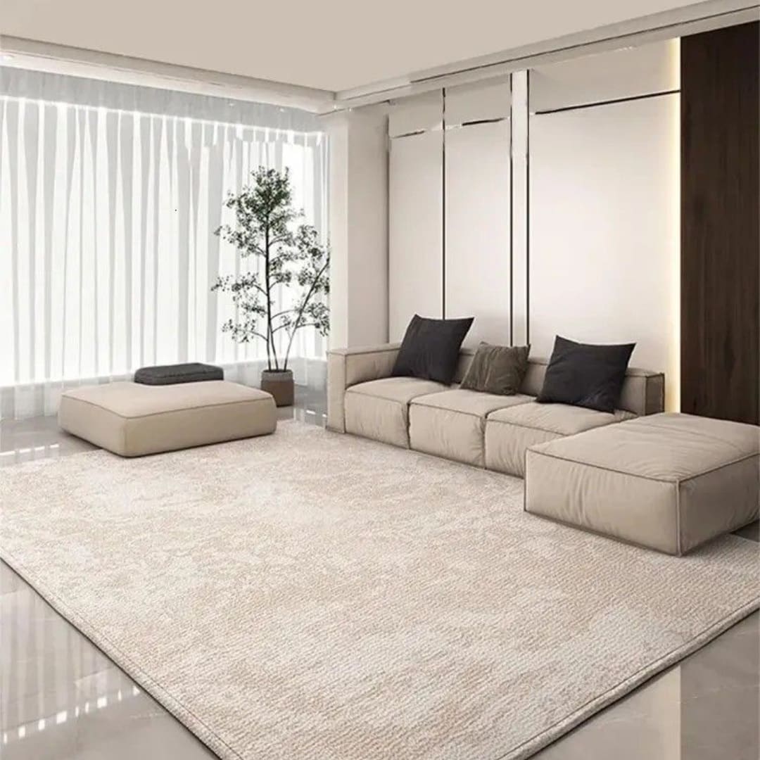 A modern living room with a beige sectional sofa, large matching ottoman, light textured rug, potted plant, and floor-to-ceiling sheer curtains covering tall windows.