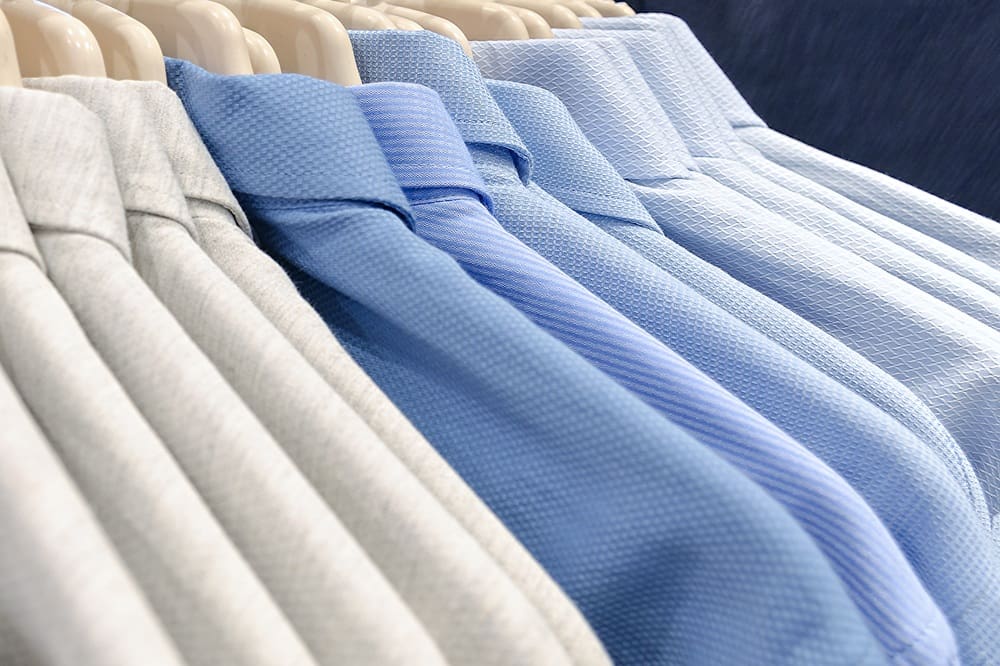 A row of neatly hung collared shirts in shades of blue and light gray on wooden hangers.