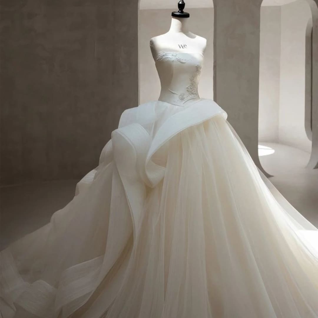 A strapless white wedding gown with a fitted bodice and voluminous, layered tulle skirt displayed on a dress form in a minimalist, light-filled room.