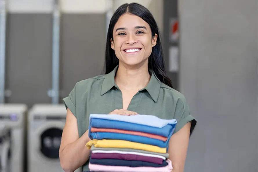A woman smiles while holding a stack of folded clothes in a laundromat.