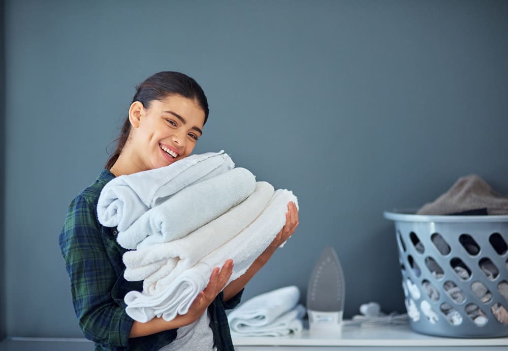 Woman smiling and holding a stack of folded towels, with a laundry basket and iron in the background.