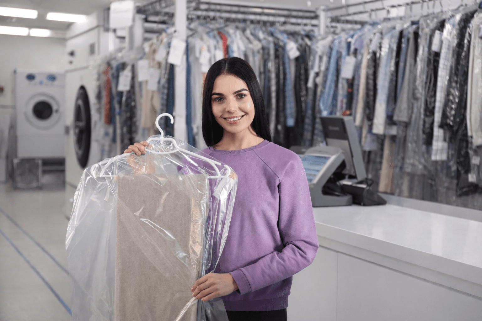 A woman in a purple sweatshirt holds dry cleaned clothes in plastic at a counter in a laundromat or dry cleaning shop. Racks of clothing and machines are visible in the background.
